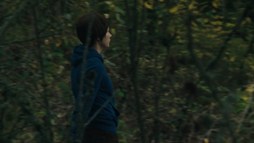A young woman wearing boyish hair and a blue hoodie walks through a shady woodland