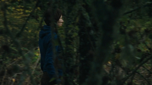 A young woman wearing boyish hair and a blue hoodie walks through a shady woodland