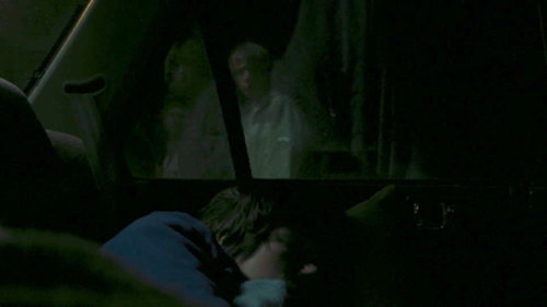 This image depicts a young woman sleeping inside a car, at night. A young man walking past peers in through the car window.