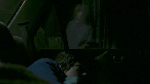 This image depicts a young woman sleeping inside a car, at night. A young man walking past peers in through the car window.
