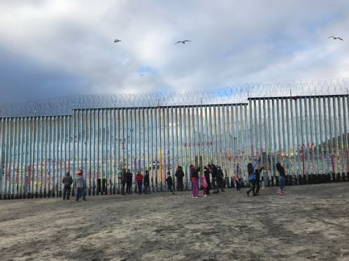 Landscape photograph taken on a beach of central American migrants framed against a metal wall dividing a national border.