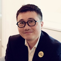 A headshot of Dr Xiaodong Lin. Dr Lin is smiling, sat in a classroom with a blackboard and bookshelf in the background.