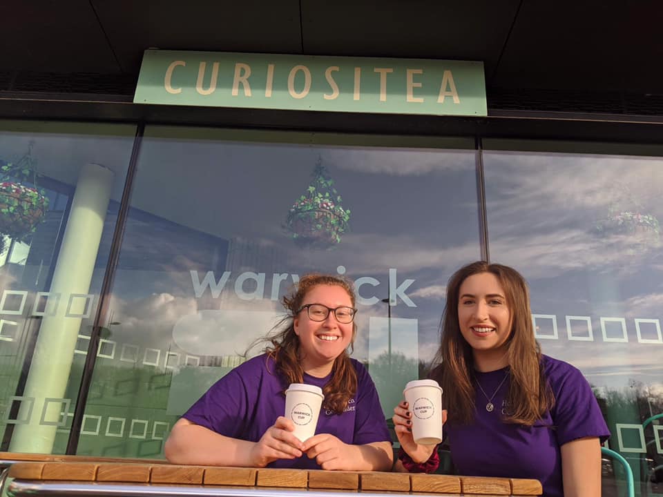 Founders of Warwick Cup, Hollie and Ellie, sitting in front of 'Curiositea' (a cafe) on campus, holding reusable cups