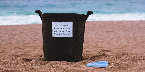 A face mask discarded on a beach, next to a bin that says 'Don't forget to protect the planet after you're done protecting yourself'