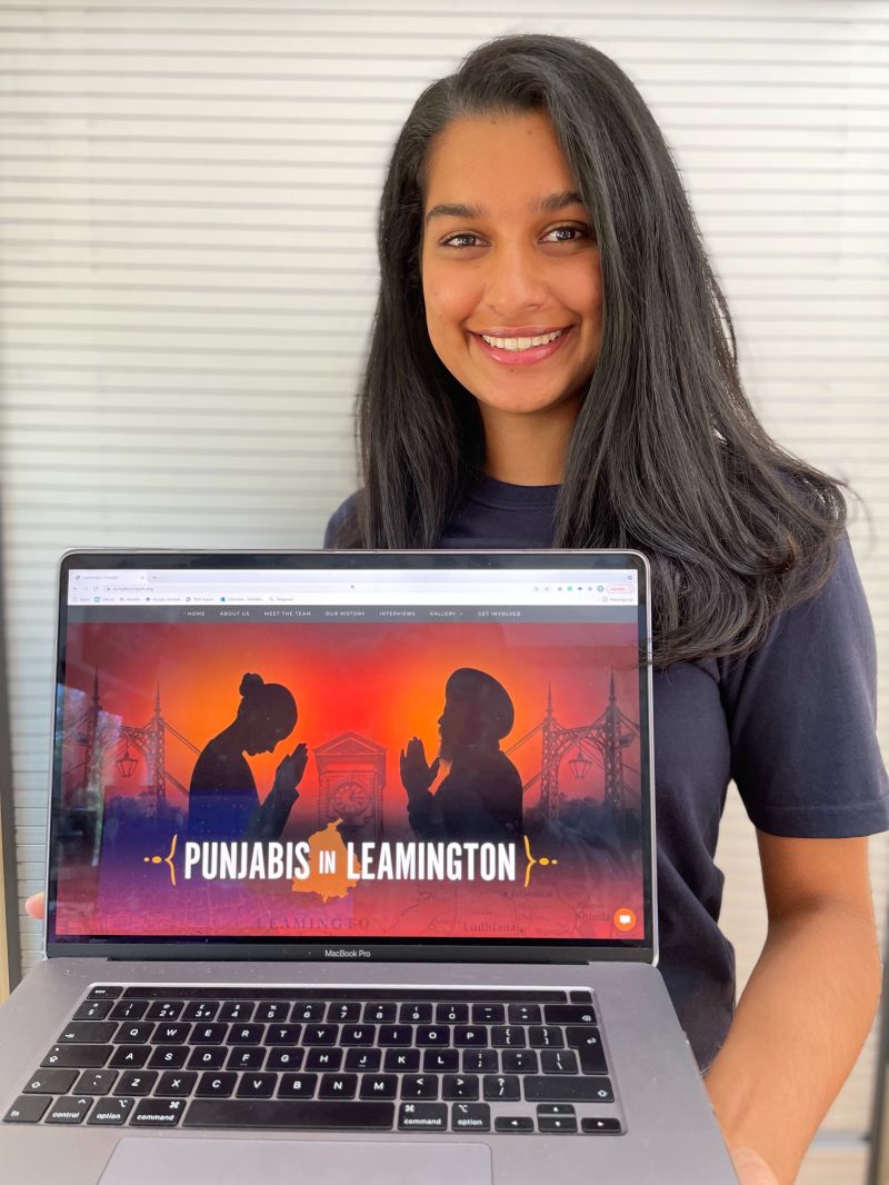 Daveena is smiling at the camera, holding a laptop which has her 'Punjabis in Leamington' website on the screen