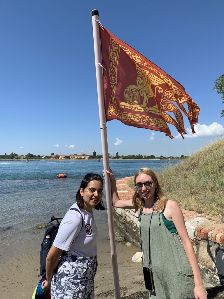 Two students standing with the Venice flag, one of them is holding onto the flag pole. The lagoon can be seen in the background.