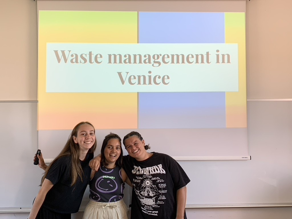 Three students stood at the front of the room with their arms around each other. They are presenting on 'Waste Management in Venice', which is the title of the presentation slide on the screen behind them.