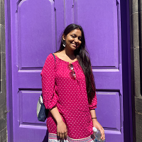 Shehzadi is featured in this photo, stood outside in front of a purple door. Shehzadi is smiling at the camera, wearing a pink dress.
