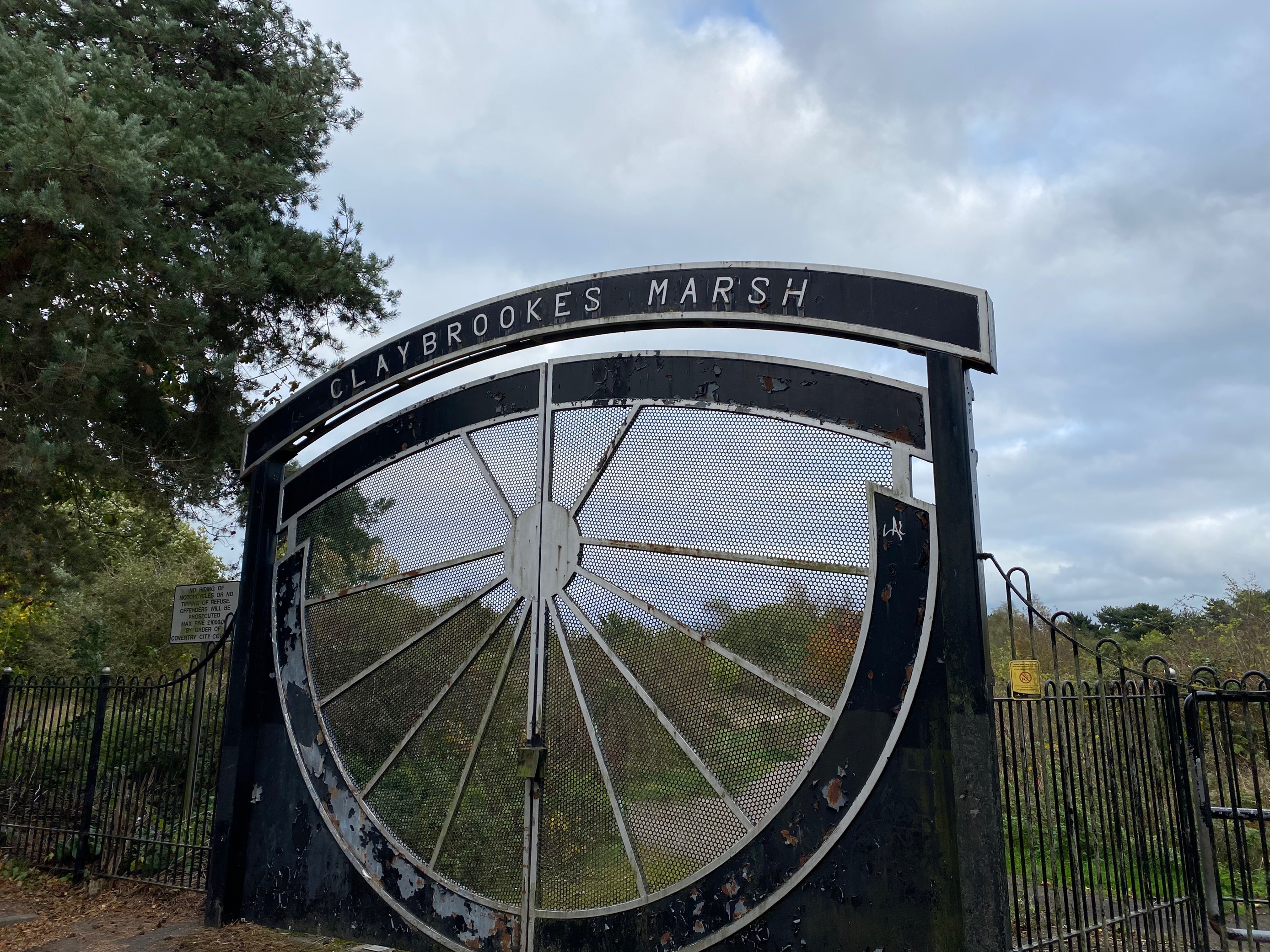 Picture showing the entrance gates to Claybrookes Marsh nature reserve. The gates are tall and made of metal, decorated with a pattern like spokes of a wheel and topped with the nature reserve's name. In the background you can see the nature reserve looking autumnal.