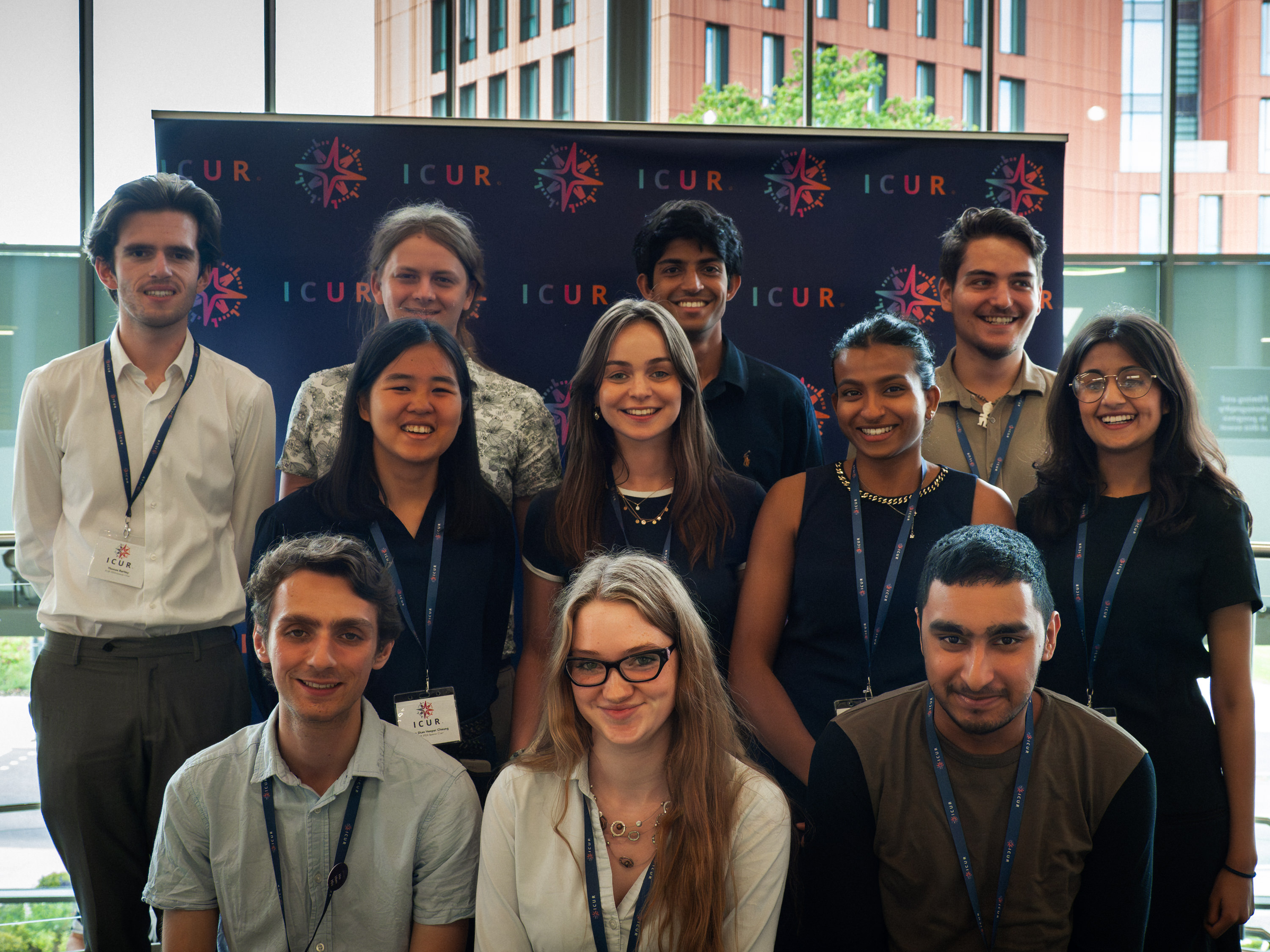 Decorative. A group of student staff wearing ICUR lanyards standing in front of a large banner displaying the ICUR logo (a compass next to ICUR text, in a gradient from turquoise, purple, pink, coral, orange).