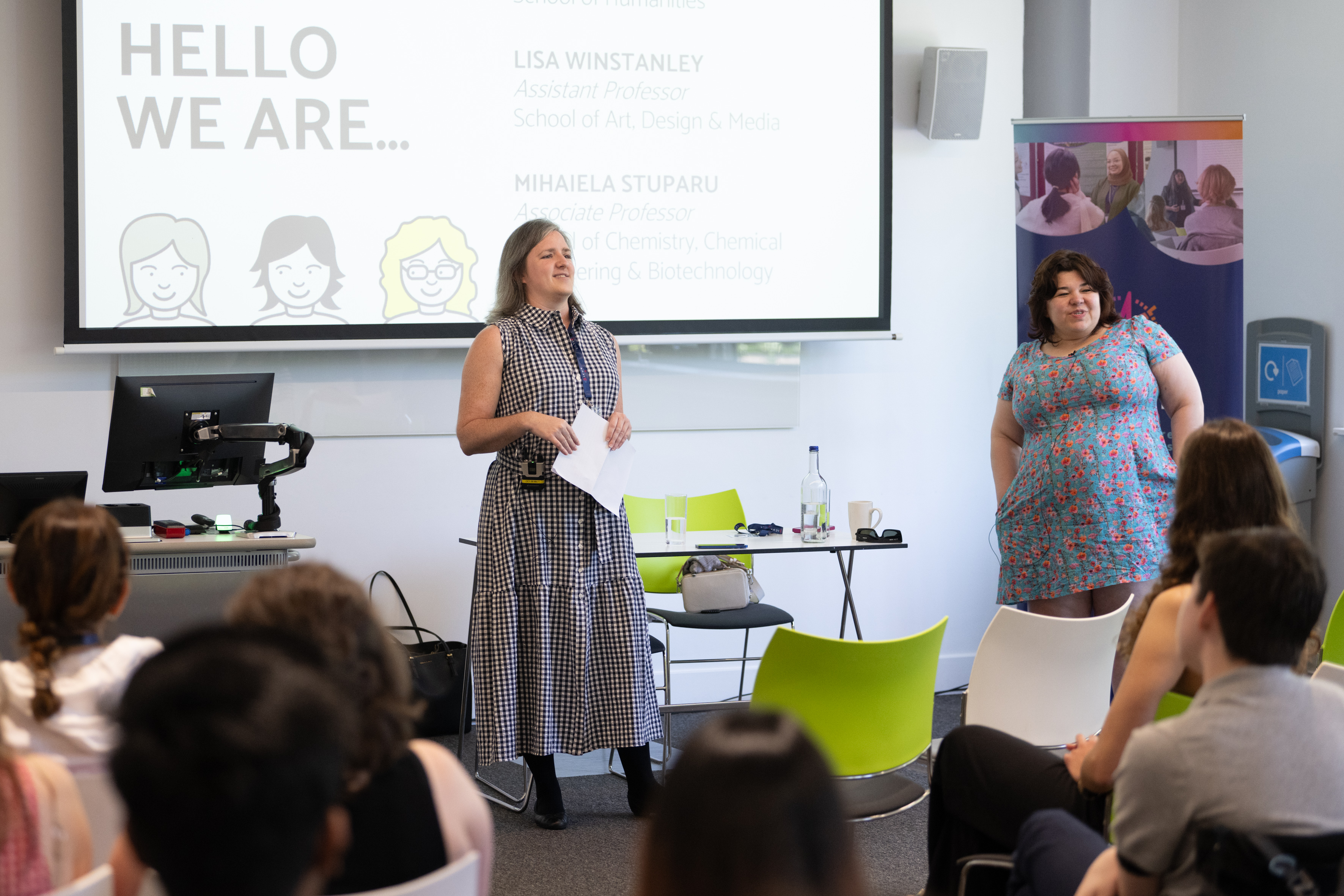 Two women are in the image. On the right, a woman with shoulder-length dark hair is wearing a blue dress. In the centre of the image, a woman with shoulder-length grey hair and wearing a black and white dress speaks into a microphone. Both women are in front of an audience.