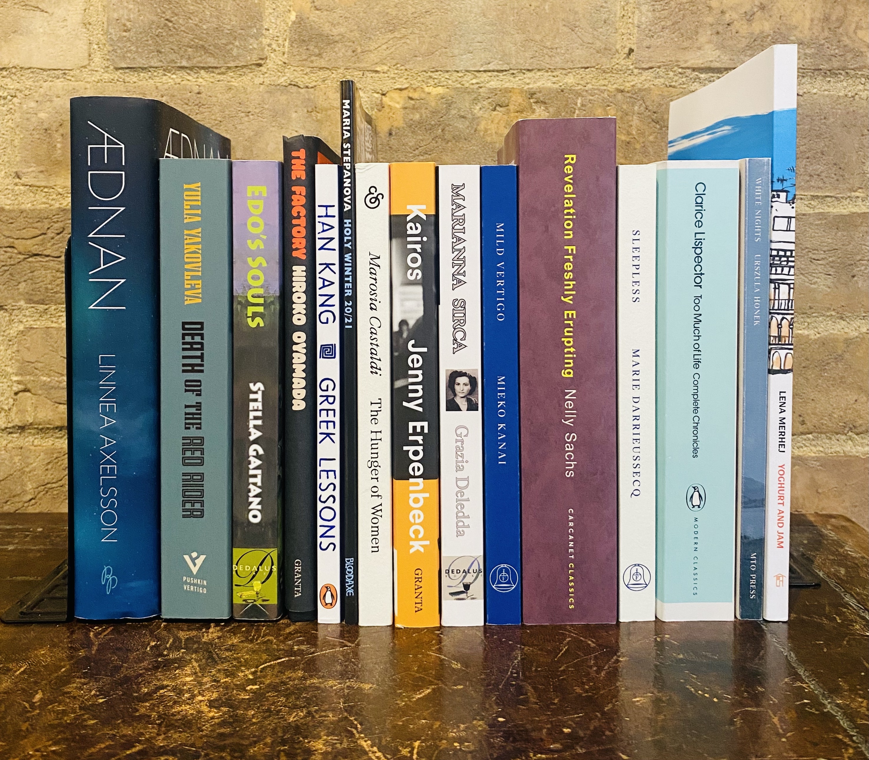  A photograph of the spines of the 15 titles longlisted for the Warwick Prize for Women in Translation in 2024. The books are stood on a wooden desk against a brick wall. 