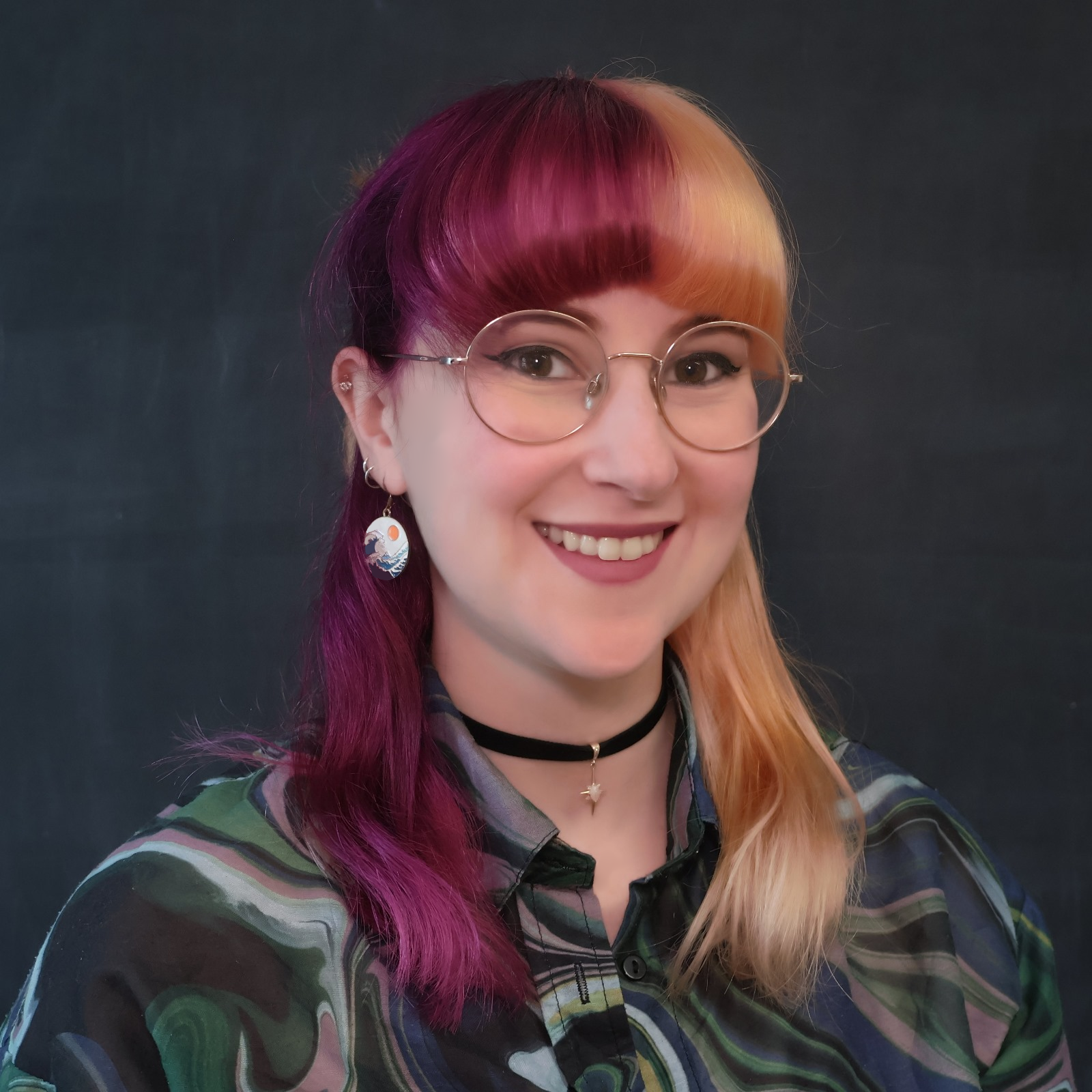 A headshot of Kat infront of a blackboard. She has purple and peach hair split dyed with a full fringe. They're wearing round framed glasses and smiling