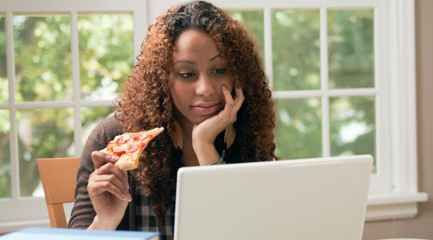 A student eating a slice and pizza whilst also looking at a laptop