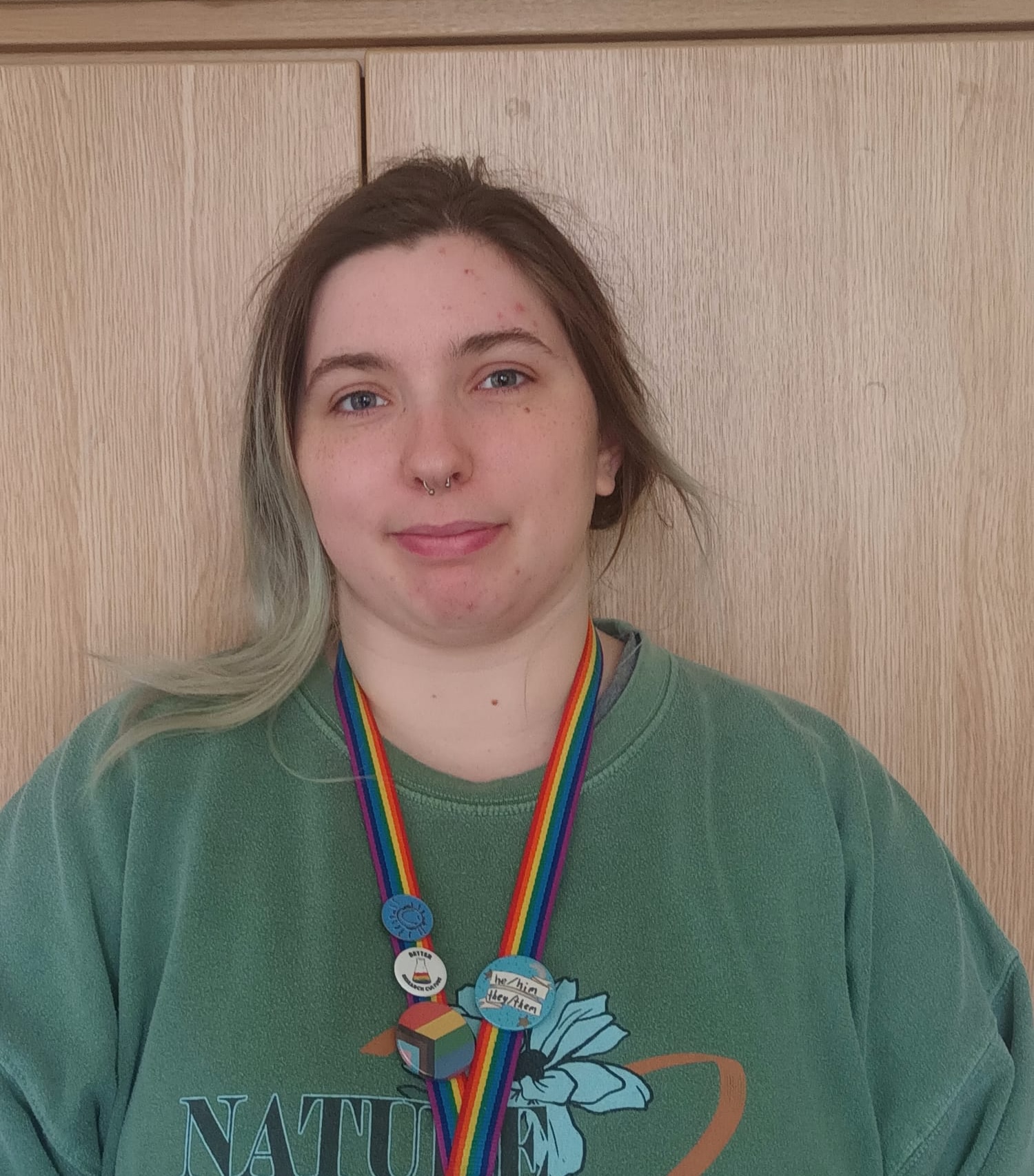 Lee is a white, femme-presenting person with long hair that has long brown roots and a faded green colour. They are wearing a dark green jumper with the word "nature" visible, and a lanyard with a rainbow pattern. The lanyard has several buttons: a blue badge with a smiley face that reads "Warwick Volunteers"; a white badge with a rainbow-coloured liquid in a conical flask that reads "Better Research Culture", a large pin with the progress flag on and a blue badge with a paper ribbon that reads "he/him, they/them". Lee is standing against a backdrop of light-coloured wood and slightly smiling at the camera.