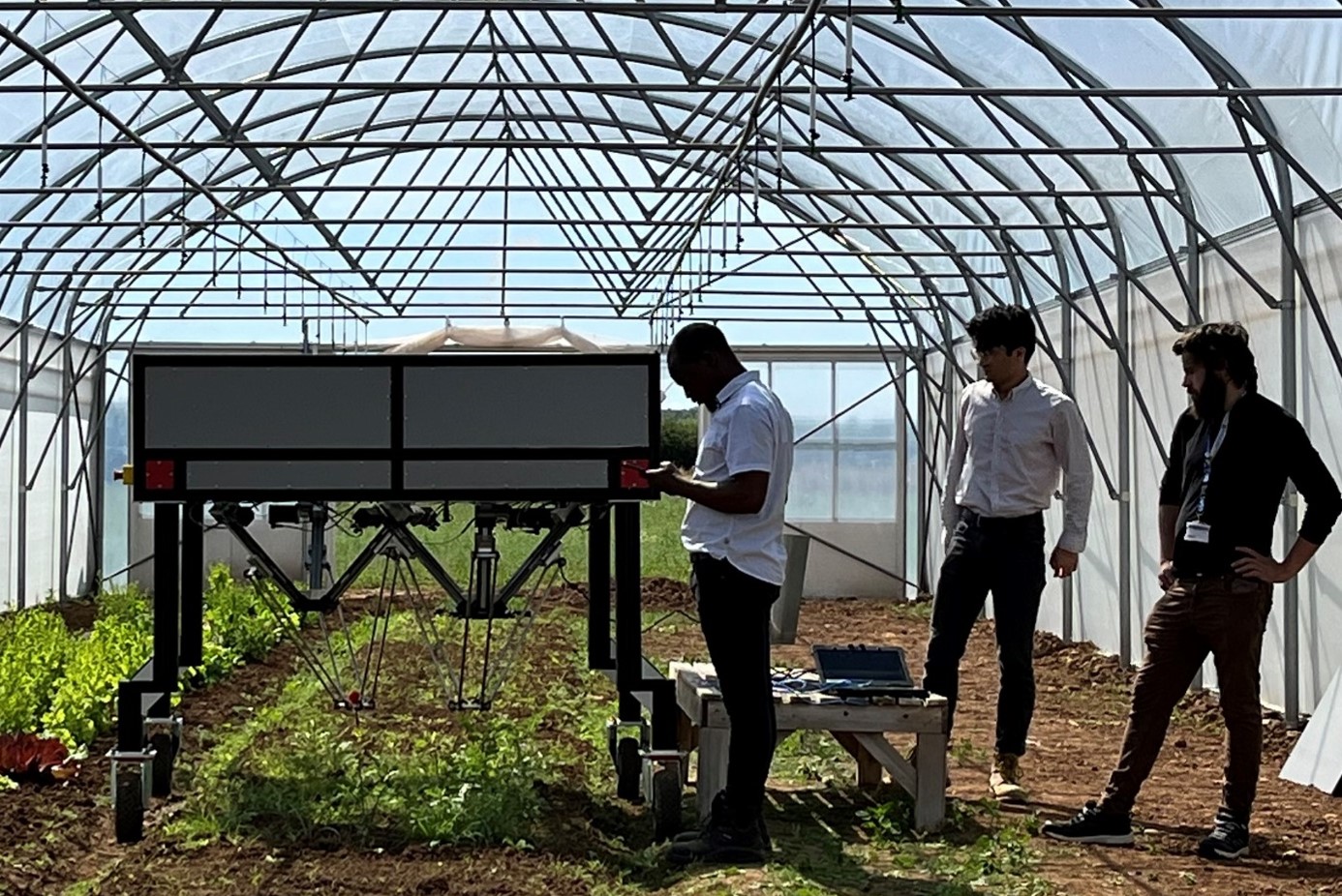 A large box on wheels stands above a row of lettuces in a poly tunnel. Three men watch on looking at computers and control panels.