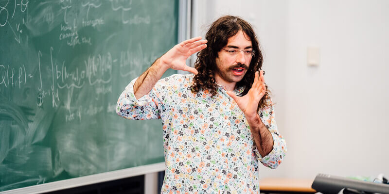 A male lecturer with long hair and a floral shirt teaching in front of a chalk board