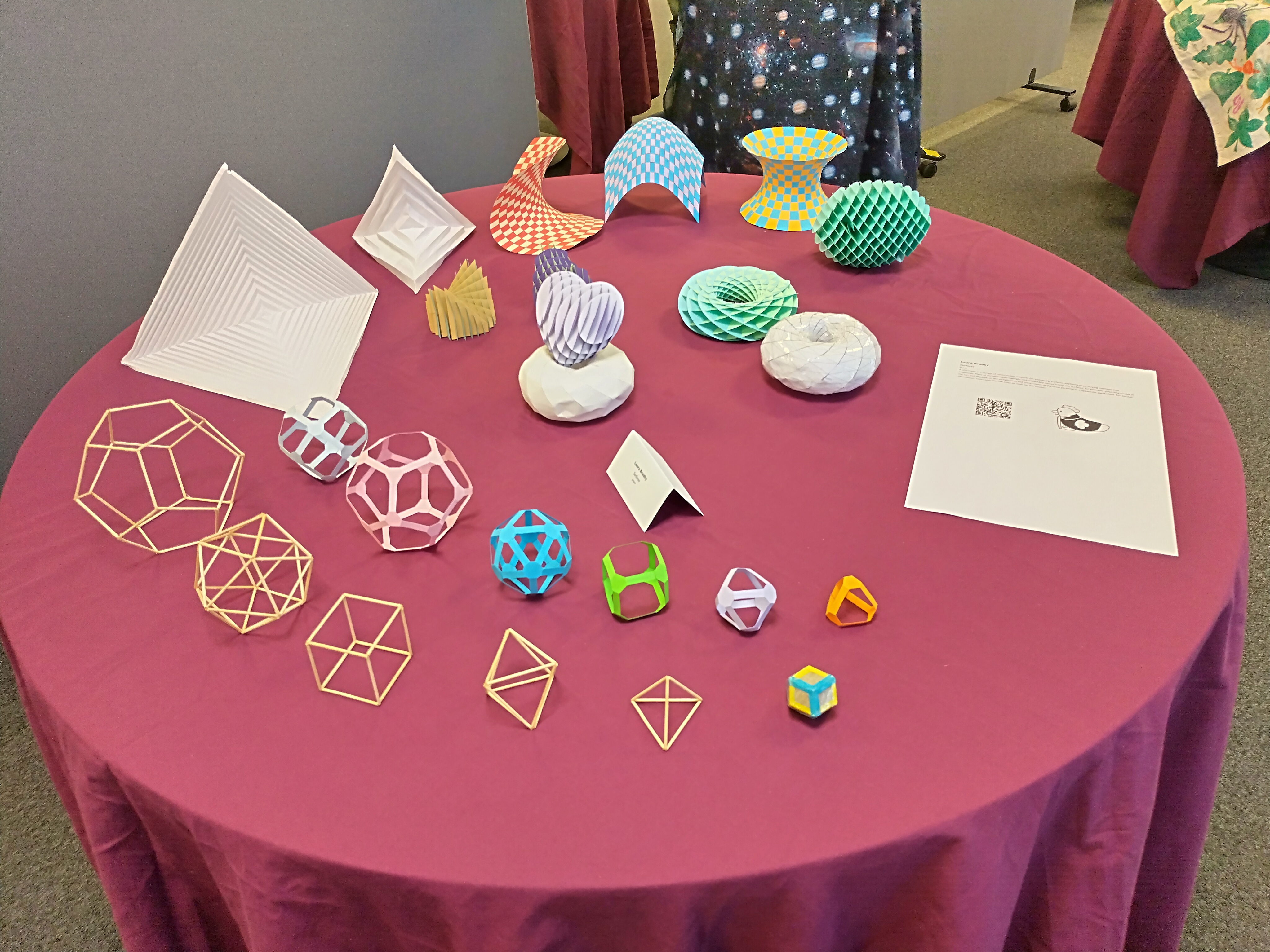 A table set up for an art exhibition, with lots of mathematical crafts, including: three different curved surfaces made by weaving strips of paper, a hyperbolic paraboloid  approximated by folding a square piece of paper, several different shapes made from pieces of card slotted together, a torus made from paper, and two sets of the platonic solids - one made from cocktail sticks, the other made from card with the faces cut out.