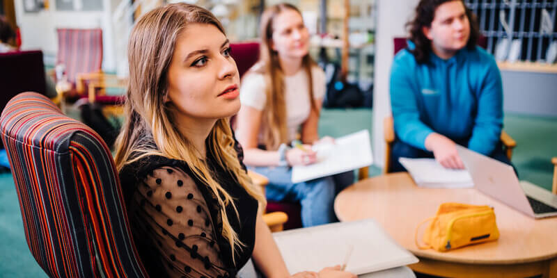 One student sitting in a chair listening to a speaker who is out of shot, with two more in the background out of sharp focus