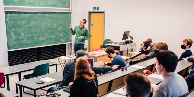 A wide shot of a classroom, with students sitting in rows facing a lecturer who is teaching at the chalk board