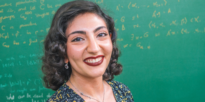 A female student smiling in front of a chalk board covered with mathematical equations