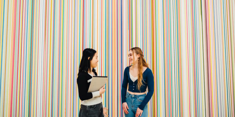 Two students laughing, while stood in front of some colourful artwork 