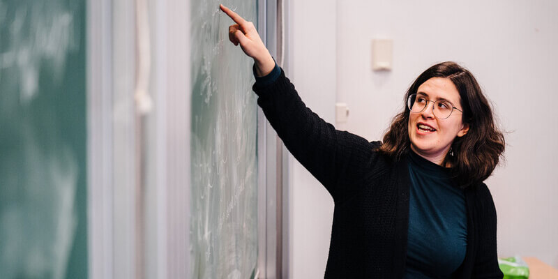 A female lecturer teaching and pointing at a chalk board