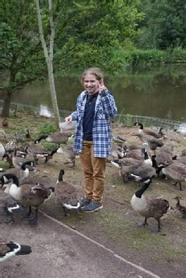 A person standing in front of the pond in Tocil woods, surrounded by geese. They are holding a bag of seeds in one hand, and posing for the camera with the other.