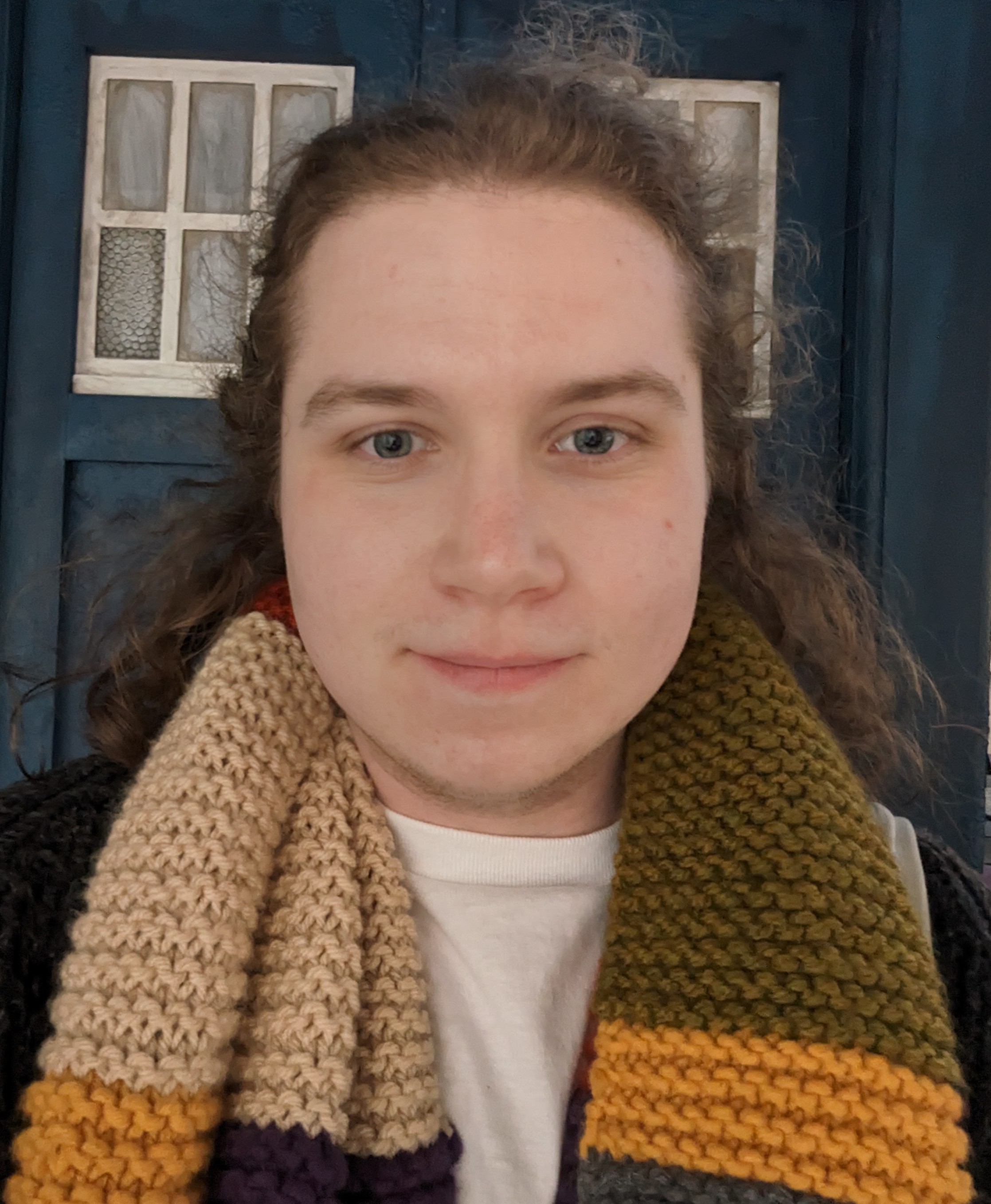 Photo of Elliot Vincent. They are framed from the shoulders up, smiling at the camera. They are wearing a colourful knitted scarf and the background is the doors of the TARDIS from Doctor Who.
