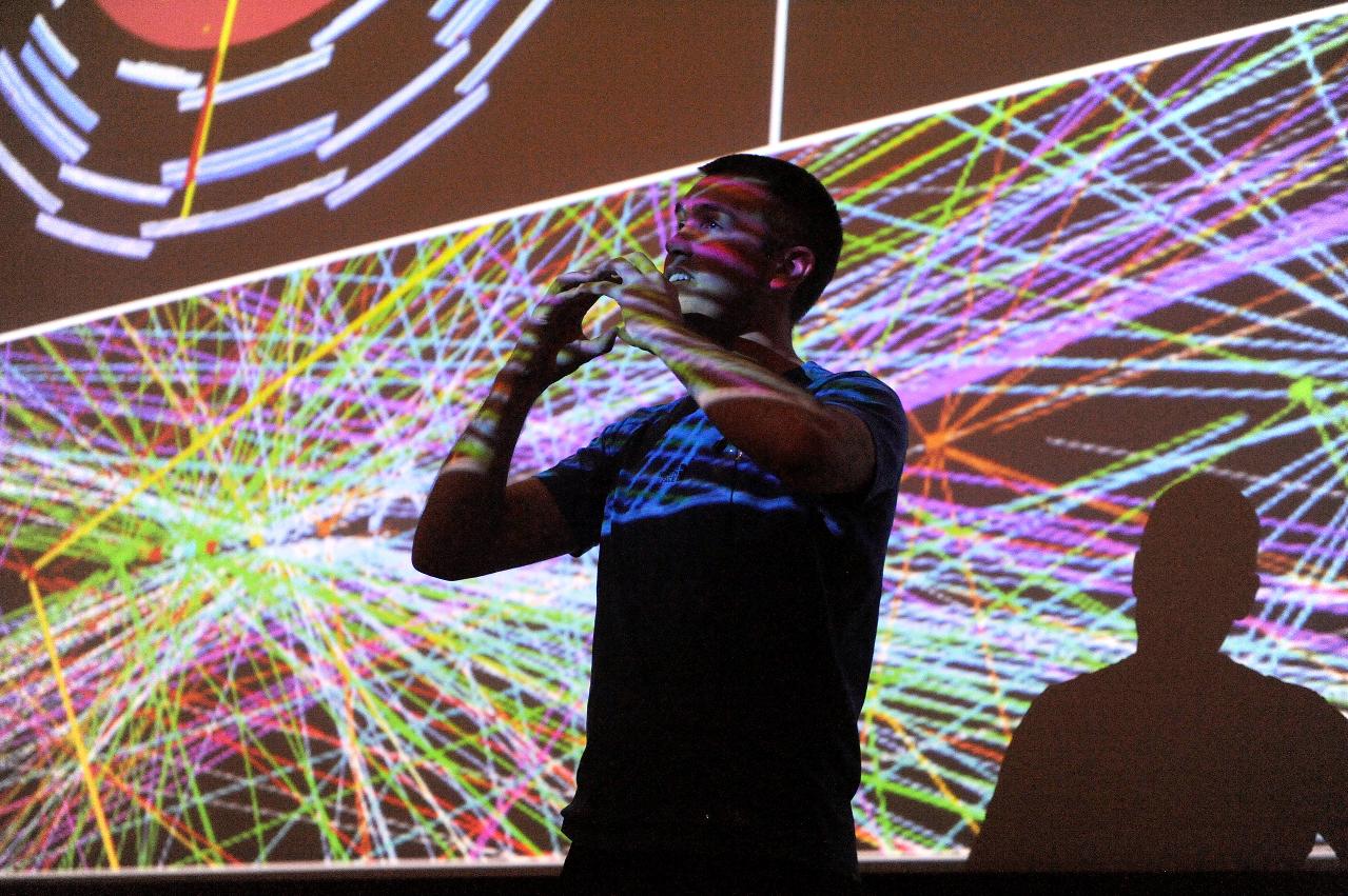 Dan Scully giving a particle physics lecture. He is only partially lit, and there is an image of particle trails being projected onto and behind him.