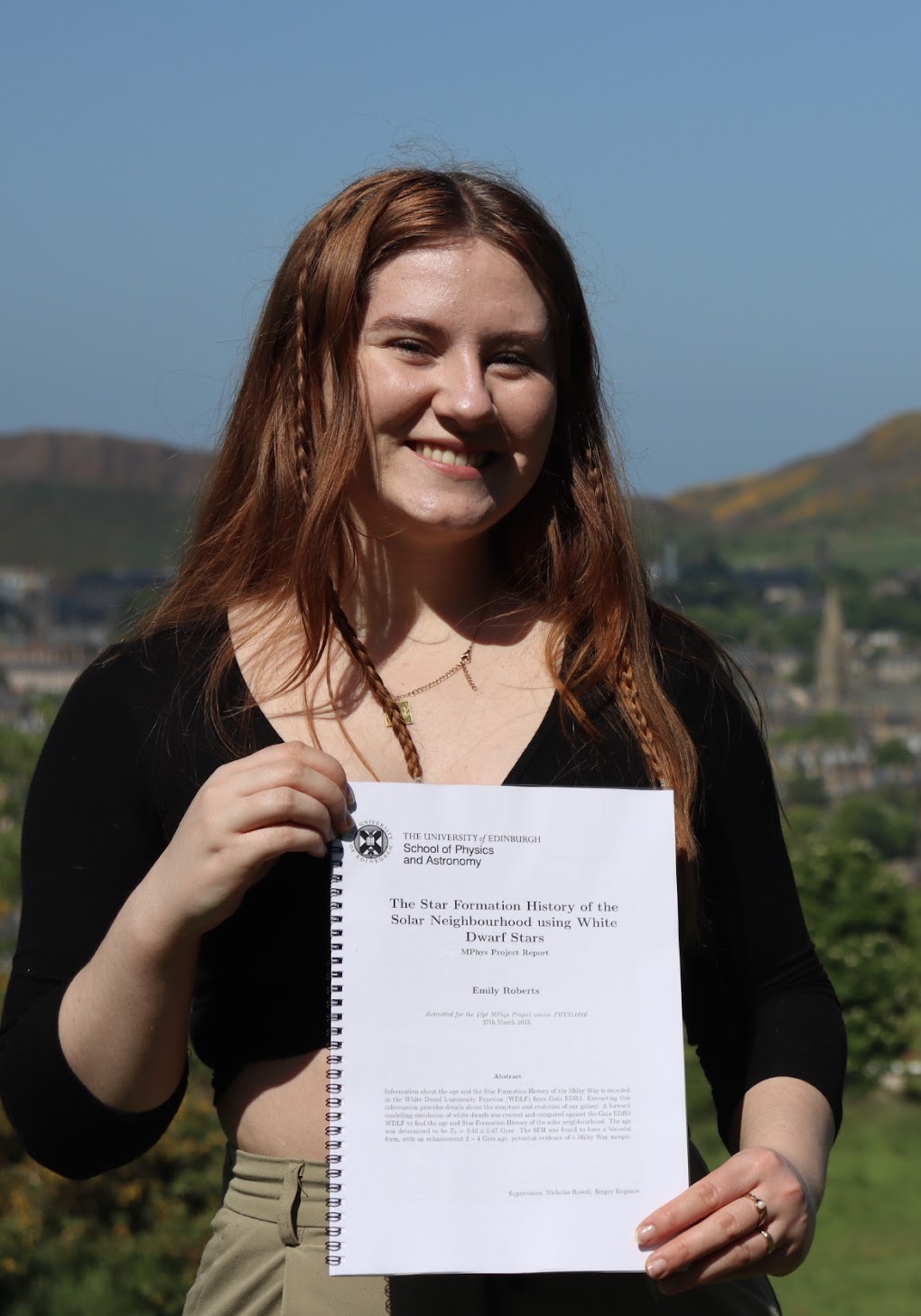 Emily (a twenty four year old woman with long red hair) is smiling while holding her dissertation. She is wearing a black top and pale green trousers.