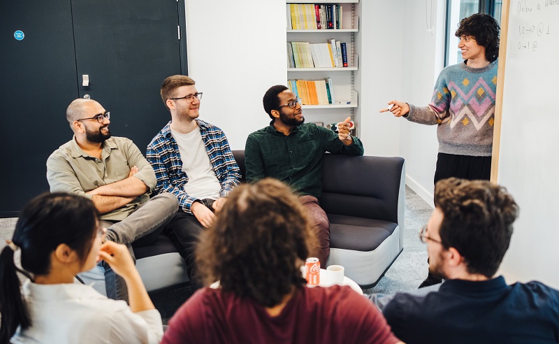 A group of students sit around a whiteboard, in discussion.
