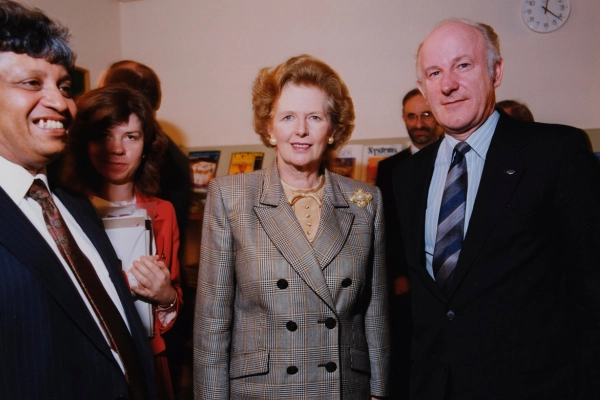 Former UK Prime Minister Margaret Thatcher smiles to the camera alongside people wearing formal business attire.