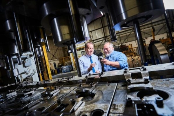 Two people stand beside large industrial machinery in a factory, examining a metal component at a work bench.