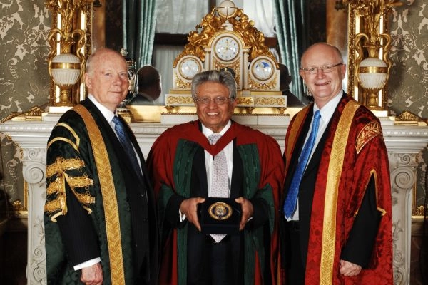Lord Bhattacharya smiles to camera whilst holding a gold medal, flanked by two people in ceremonial robes