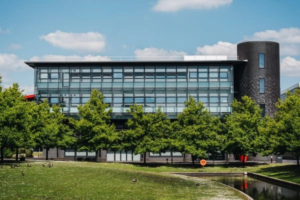 A modern, glass-fronted building with a cylindrical tower sits behind a lake on campus