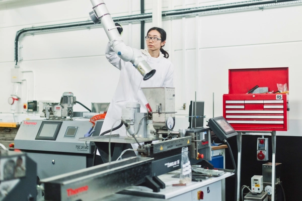 Person wearing a lab coat operating a machine in a laboratory, surrounded by industrial equipment, control panels, and a red tool cabinet.