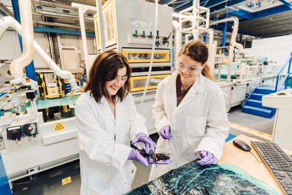 Two people wearing lab coats and safety glasses examine a small material sample in an industrial laboratory, with manufacturing equipment and machinery in the background