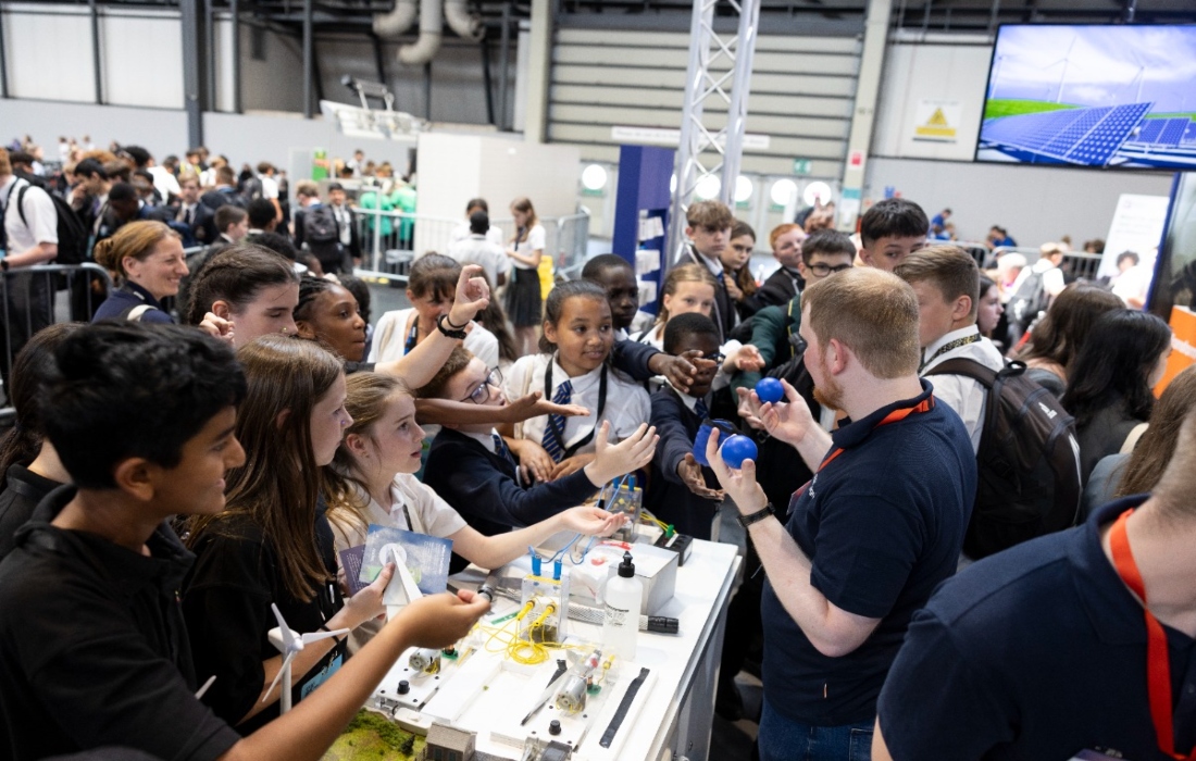 Children at a science fair interacting with engineers holding examples of manufactured products.