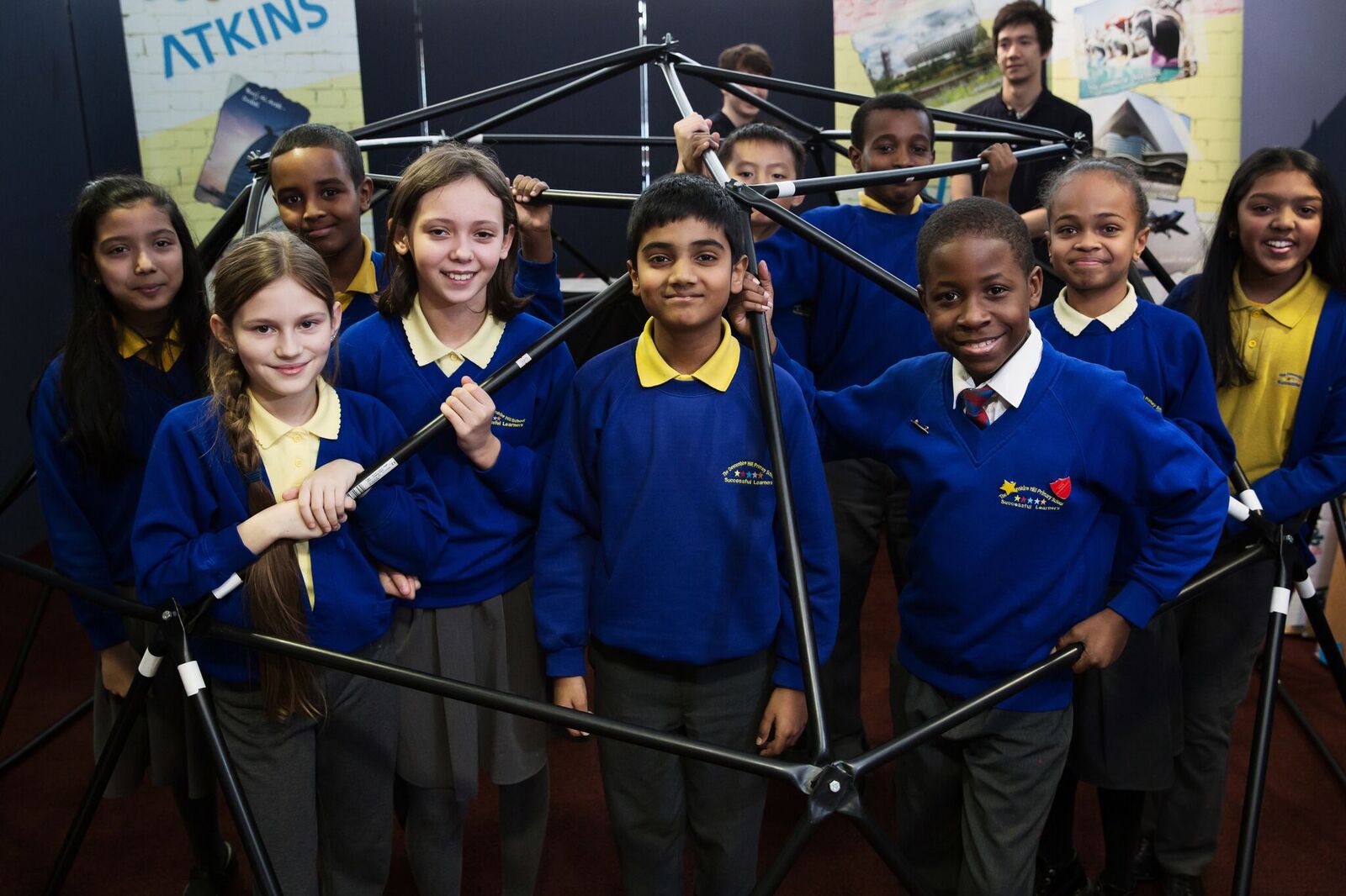 Students holding a metal framework that is shaped like a hexagon. It is unclear what the particular component is, but it is large enough for several of the students to stand inside it.