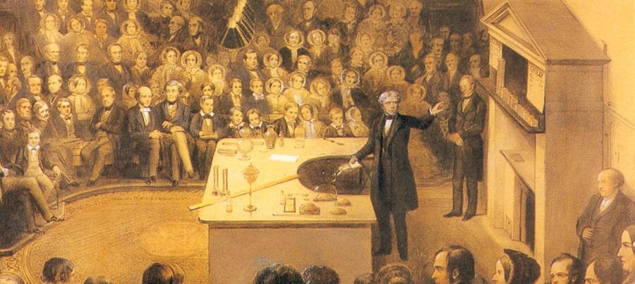 A painting of Michael Faraday presenting a Christmas Lecture. There are props and scientific instruments on a bench in front of him, while he points to objects on a shelf behind.