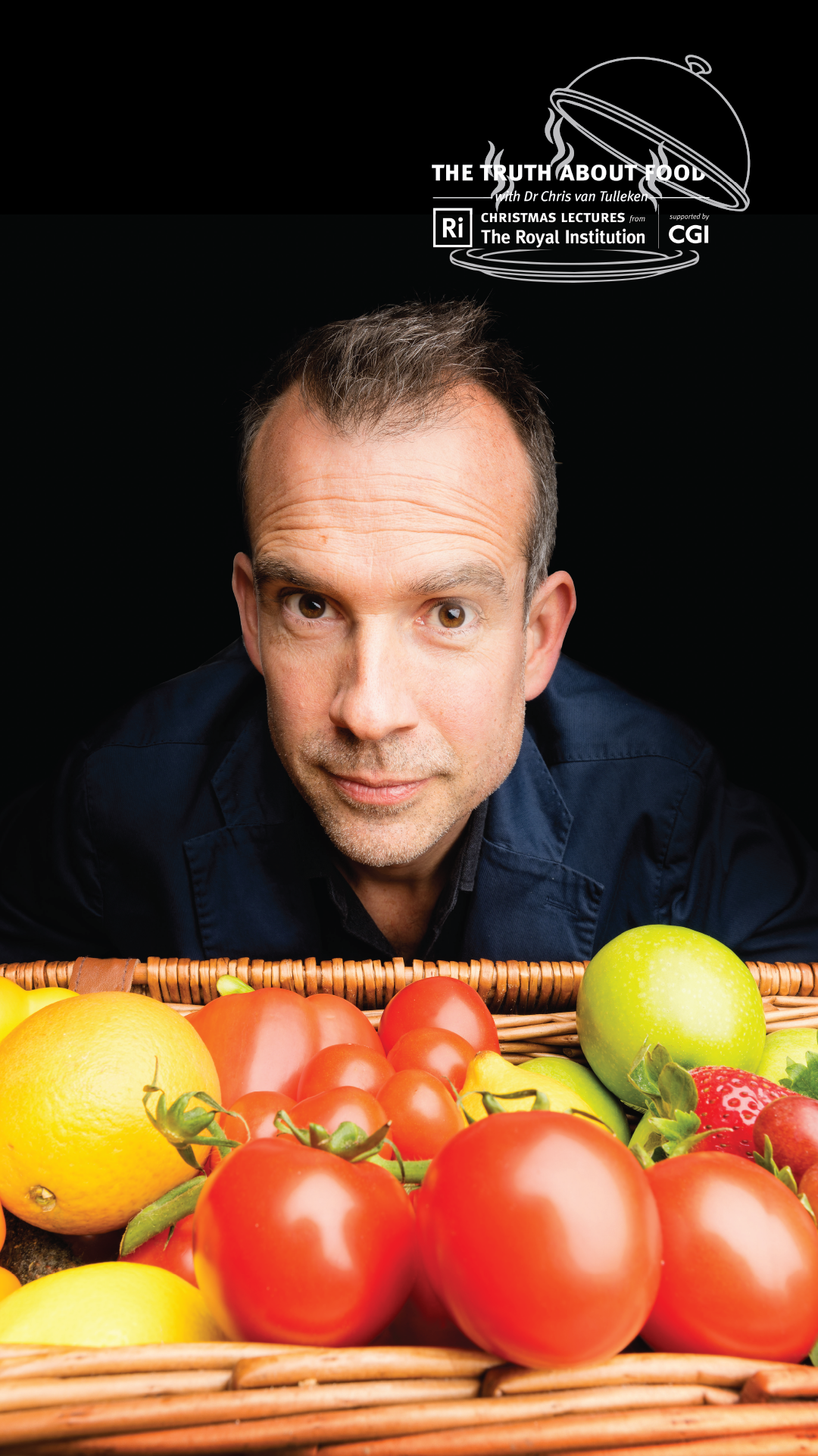 An image of Chris van Tulleken against a black backdrop. The foreground is taken up by a wicker basket of fruit and vegetables.