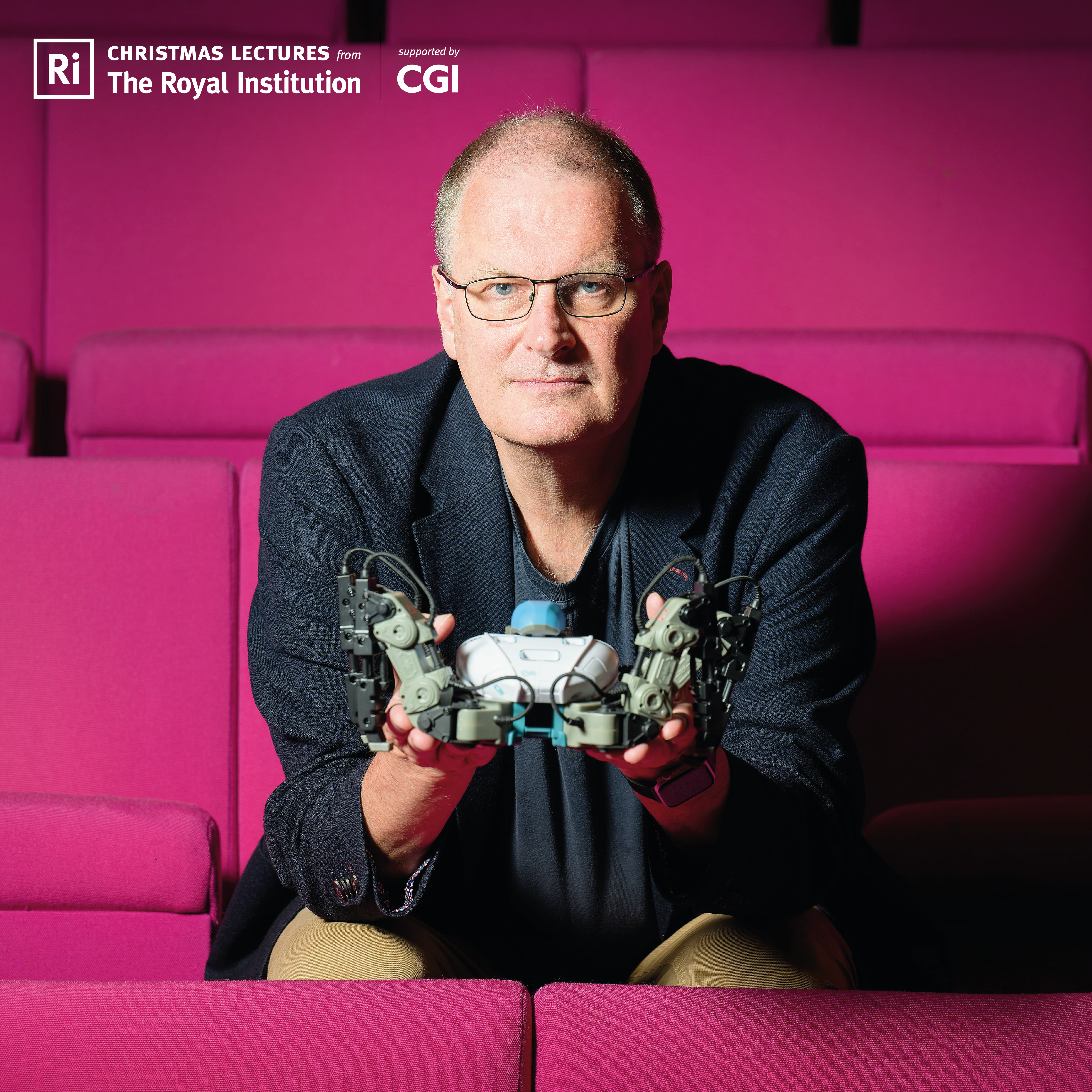 A photo of Mike Woolridge sitting on a pink lecture theatre chair. He is holding a robotic device. It is hard to see what the device is, exactly.