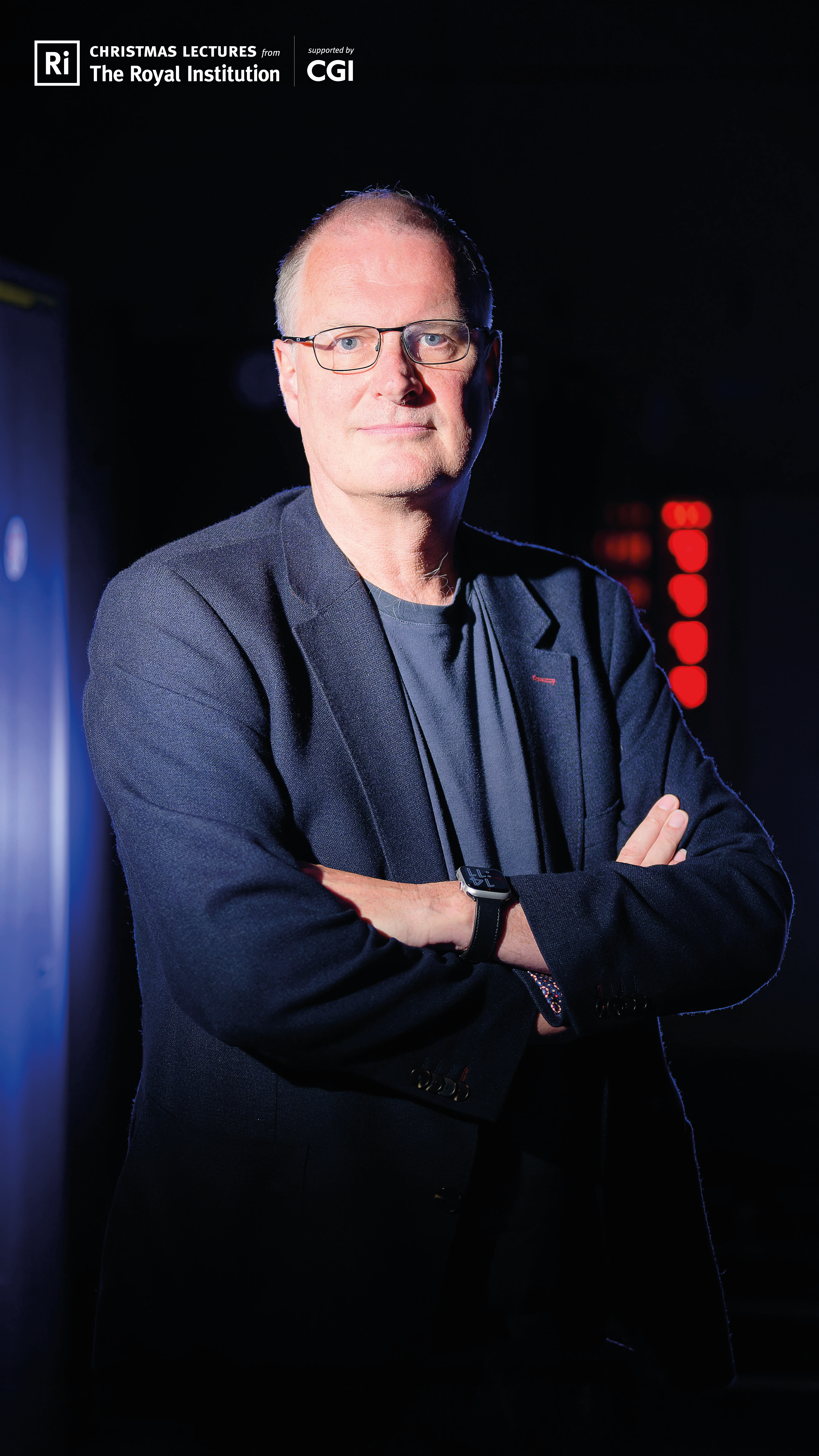 A picture of Mike Woolridge standing in front of a dark background. He is dramatically lit from one side. There are blue and red lights in the background.