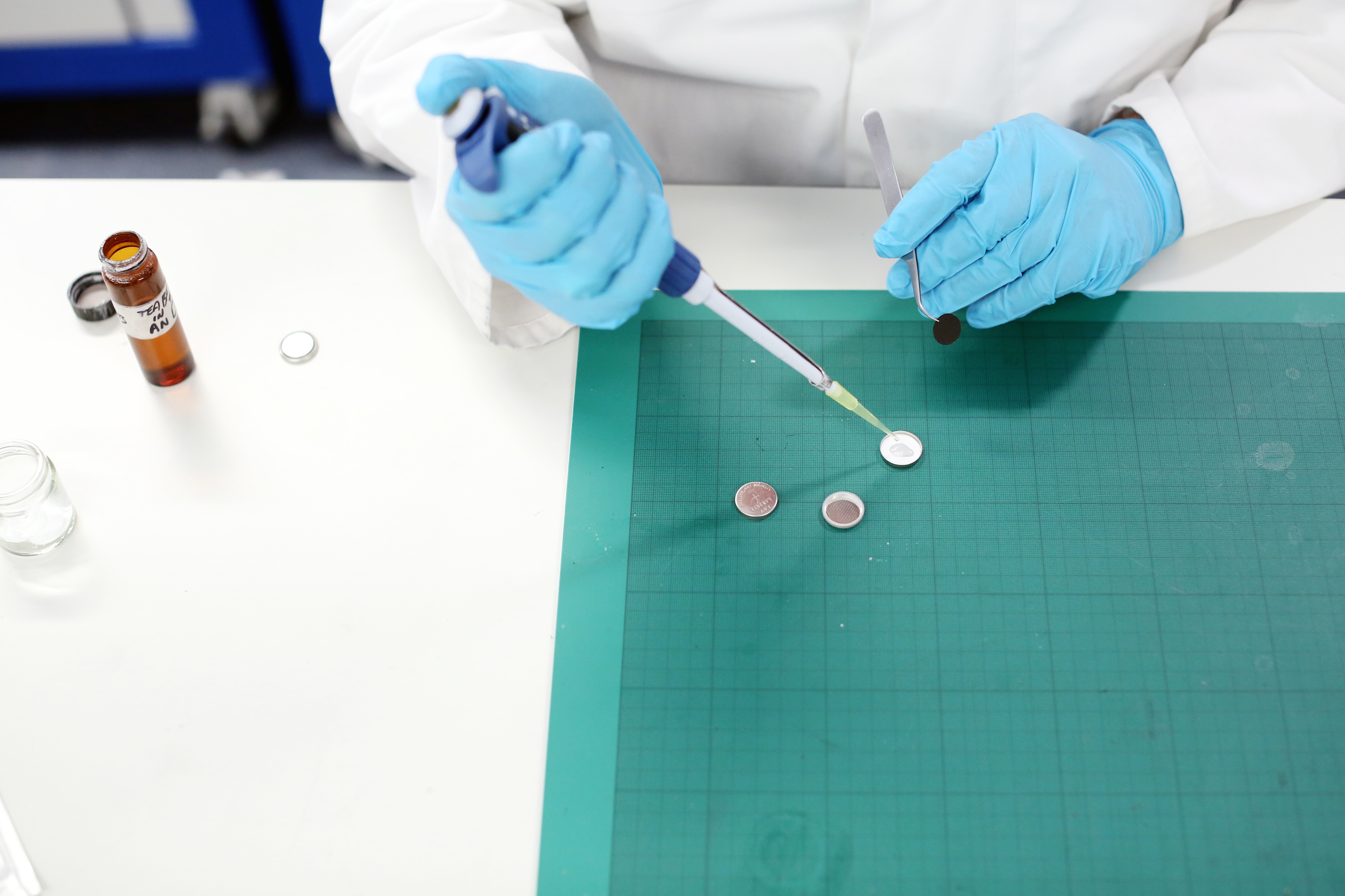 A person wearing blue lab gloves using a pipette to add liquid to a coin cell battery in a lab environment.