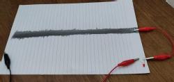 A thick, dark pencil band has been drawn onto a piece of paper. One crocodile clip is connected to the pencil line. Another sits loose nearby.