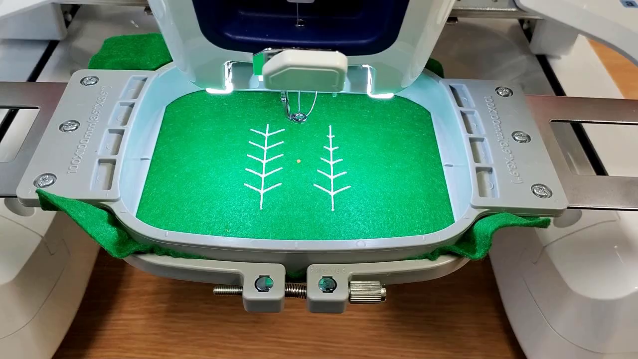 There is a digital embroidery machine with two example tree patterns stitched onto green felt.