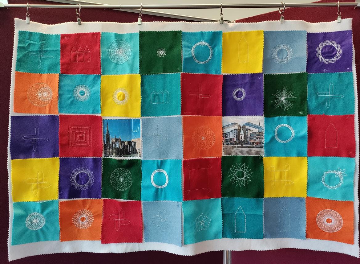 Squares of red, orange, yellow, green, purple and blue felt with white embroidery stuck onto a large rectangle of white felt. There are two square photos near the centre, of Coventry landmarks. 