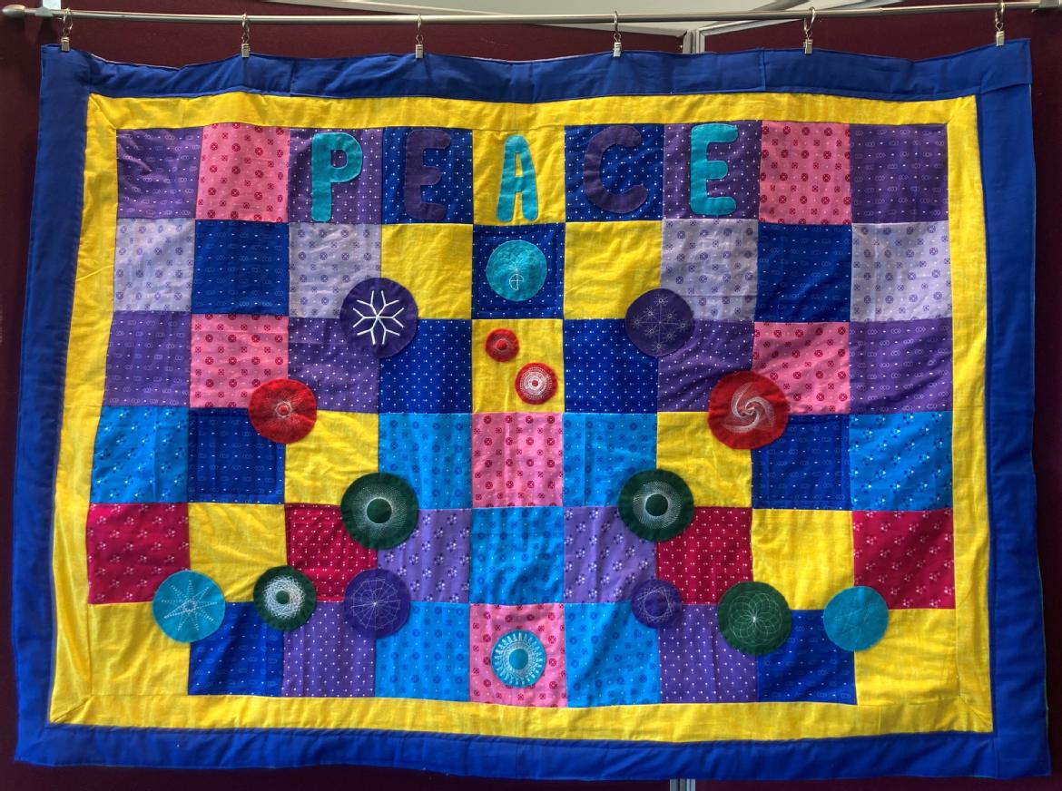 A quilt made of purple, yellow, pink and blue squares of fabric, with a blue and yellow border. The quilt says peace at the top. Attached to the quilt are felt fabric circles with white embroidery on them. 