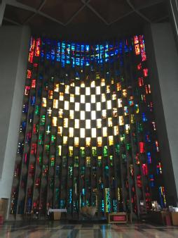 The baptistry window at Coventry Cathedral.  In the centre there is a lot of white and yellow stained glass, round the outside are blue red and green stained glass.